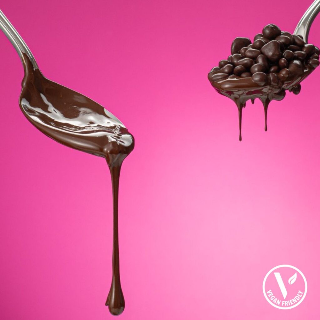vegan chocolate in a spoon and vegan chocolate coated nibs in another spoon against a pink background